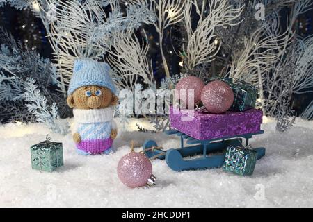 Un gnome giocattolo lavorato a maglia si erge sulla neve artificiale e una slitta giocattolo con regali si trova accanto a lui sullo sfondo di un albero artificiale bianco e blu Foto Stock