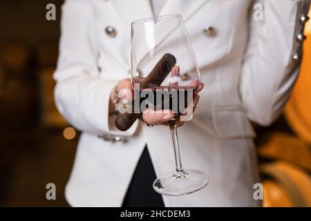 Sezione centrale di una donna caucasica sconosciuta che tiene in mano un bicchiere di vino rosso e un sigaro mentre si alza in piedi al coperto vista frontale copia spazio drink e celebratio Foto Stock