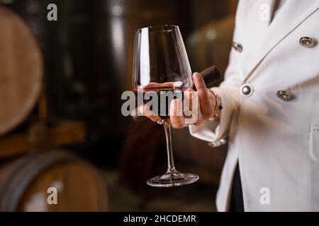 Sezione centrale di una donna caucasica sconosciuta che tiene in mano un bicchiere di vino rosso e un sigaro mentre si trova al coperto in cantina vista laterale copia spazio drink e c. Foto Stock