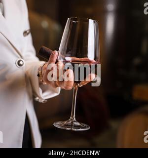 Sezione centrale di una donna caucasica sconosciuta che tiene in mano un bicchiere di vino rosso e un sigaro mentre si trova al coperto in cantina vista laterale copia spazio drink e c. Foto Stock