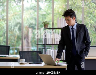 Il giovane uomo d'affari asiatico in abito nero usa il computer portatile per controllare la posta elettronica prima di uscire a pranzo pausa. Foto Stock