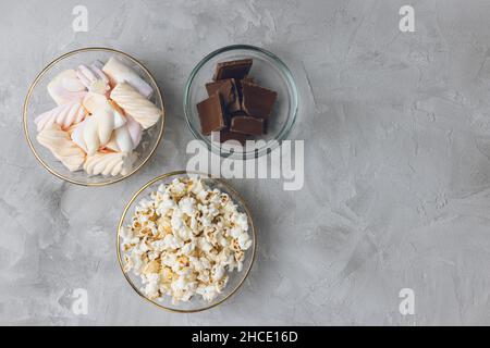 Popcorn, marshmallow pastello colorato, pezzi di barretta di cioccolato al latte rotto in ciotole di vetro su sfondo grigio. Ingredienti per cucinare dessert e dolci malsani. Vista dall'alto, spazio di copia Foto Stock