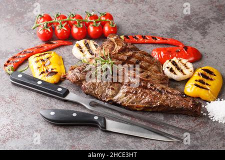 Grigliate la bistecca di manzo con osso a T con aglio, peperoni e pomodori da vicino su uno sfondo grigio di cemento. Orizzontale Foto Stock