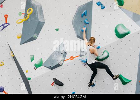 Vista posteriore del corpo di alpinista femminile attiva in abbigliamento sportivo su parete da arrampicata artificiale con manici colorati durante l'allenamento in palestra moderna Foto Stock