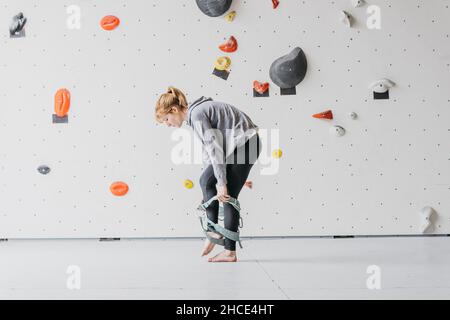 Vista laterale completa del corpo del climber femminile a piedi nudi sportivo che mette sul belay di sicurezza vicino alla roccia di arrampicata artificiale prima di allenarsi in palestra Foto Stock