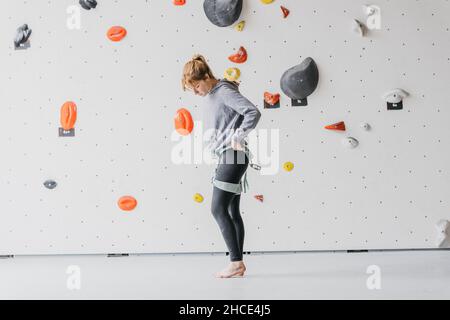 Vista laterale completa del corpo del climber femminile a piedi nudi sportivo che mette sul belay di sicurezza vicino alla roccia di arrampicata artificiale prima di allenarsi in palestra Foto Stock