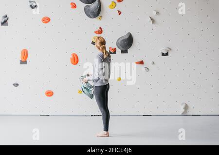 Vista laterale completa del corpo del climber femminile a piedi nudi sportivo che mette sul belay di sicurezza vicino alla roccia di arrampicata artificiale prima di allenarsi in palestra Foto Stock