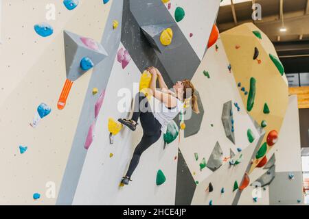 Vista laterale completa del corpo di alpinista femminile attiva in abbigliamento sportivo su parete da arrampicata artificiale con manici colorati durante l'allenamento in palestra moderna Foto Stock