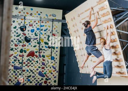 Vista laterale del corpo di sportivi anonimi a piedi nudi in abbigliamento sportivo che pratica l'arrampicata su parete con impugnature in legno in palestra leggera Foto Stock