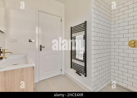 Armadietto con lavandino bianco posto contro la parete con radiatore nero in luce bagno moderno con porta chiusa e muro di mattoni bianchi Foto Stock
