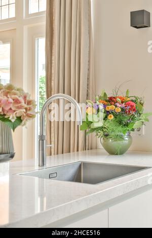 Primo piano del rubinetto nel lavandino costruito in pietra contatore con fiori colorati in fiore in cucina moderna con finestre Foto Stock