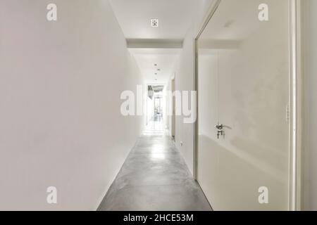 Lungo corridoio stretto con pareti bianche chiare e porte chiuse che conducono a una grande finestra di vetro in un moderno appartamento luminoso Foto Stock