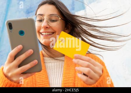 Da sotto di ragazza allegra in occhiali e cardigan arancione lavorato a maglia a pagamento per l'acquisto online con carta di plastica su smartphone Foto Stock