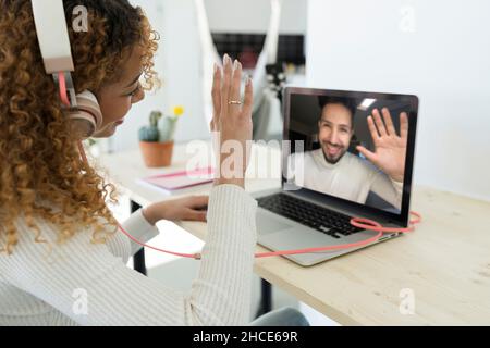Taglia la donna anonima nera con capelli ricci indossando le cuffie e seduto al tavolo mentre parli alla videochiamata con il collega maschio in ufficio Foto Stock