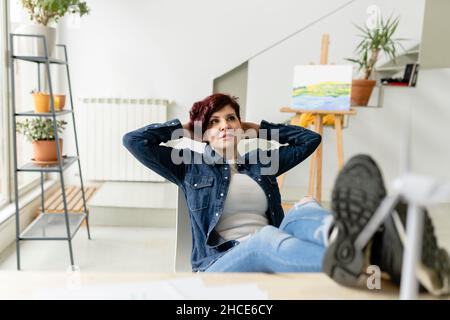 Corpo pieno di donna rilassata guardando in su con le mani dietro la testa mentre si siede nella stanza luminosa con pittura sul cavalletto a casa Foto Stock