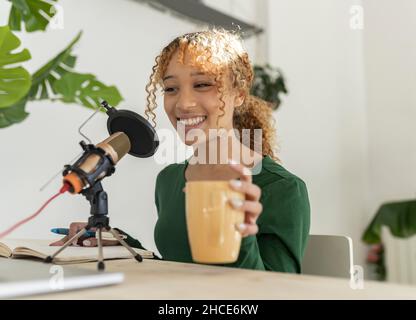 Sorridente giovane etnia femminile di registrazione podcast su laptop con microfono professionale, seduto al tavolo con un bicchiere di fresco frullato e notebook Foto Stock