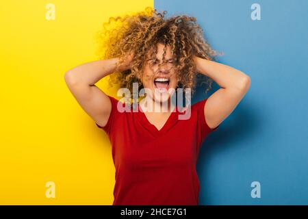Giovane donna in t-shirt rossa con espressione emozionata su sfondo giallo e blu che afferra i capelli Foto Stock