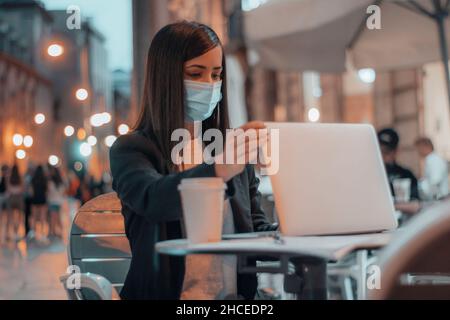 Donna d'affari con maschera chirurgica lavoro di finitura chiusura del suo computer portatile seduta in una caffetteria bere caffè di notte Foto Stock