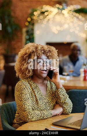 Felice donna d'affari africana in occhiali ridendo mentre si fa una conversazione sul telefono cellulare seduto al tavolo in caffetteria Foto Stock