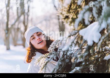Giovane donna sorridente curly cammina nel parco invernale e nascondersi dietro l'albero di Natale coperto di neve. Emozioni positive, freschezza, vacanze. Foto Stock