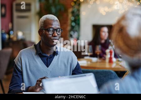 Uomo d'affari africano in occhiali seduti al tavolo in caffè, ha un incontro d'affari con i suoi colleghi Foto Stock