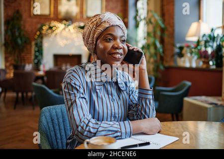 Elegante donna d'affari africana che ha una conversazione sul telefono cellulare, mentre fa appunti in notebook e bere caffè al tavolo in caffè Foto Stock