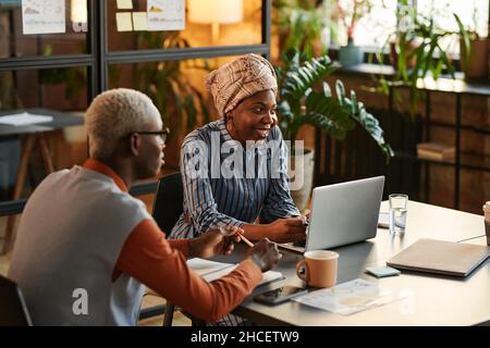 Donna d'affari africana sorridendo e parlando con i suoi colleghi mentre siede al tavolo durante la riunione, lavora in squadra con i suoi colleghi Foto Stock