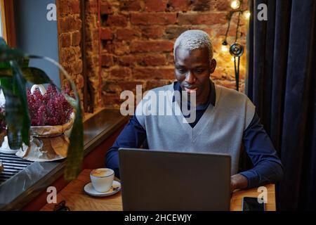 Giovane africano che lavora online su un portatile e beve caffè mentre si siede al tavolo in un bar Foto Stock