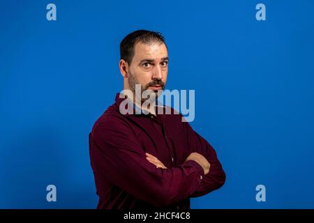 Ritratto di serio e preoccupato spagnolo barbuto uomo con le braccia incrociate, isolato su sfondo blu studio Foto Stock