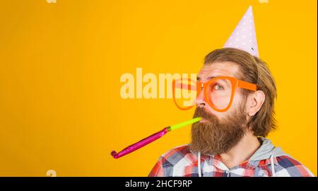 Uomo bearded nel cappello di partito soffia noisemaker. Ragazzo felice in bicchieri grandi in berretto cono di compleanno. Spazio di copia. Foto Stock