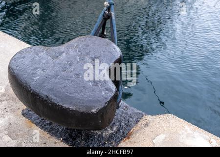 Corda di ormeggio della nave intorno ad un bullard del porto sul molo del porto, sfondo ondulato dell'acqua di mare, spazio di copia. Palo in metallo nero ancorato al pavimento in cemento della banchina Foto Stock