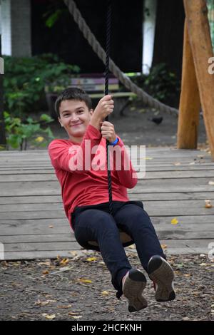 Giovane ragazzo attraente scende su una zipline in un parco divertimenti e sorride Foto Stock