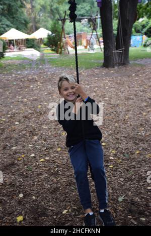 Giovane ragazzo bianco attraente scende su una zipline in un parco divertimenti e sorride felicemente Foto Stock