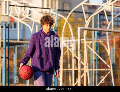 Adolescente carino in felpa con cappuccio viola che gioca a basket. Ragazzo giovane con palla imparare dribble e sparare sul campo della città. Hobby per bambini, Foto Stock