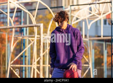 Adolescente carino in felpa con cappuccio viola che gioca a basket. Ragazzo giovane con palla imparare dribble e sparare sul campo della città. Hobby per bambini, Foto Stock