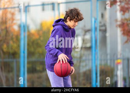 Adolescente carino in felpa con cappuccio viola che gioca a basket. Ragazzo giovane con palla imparare dribble e sparare sul campo della città. Hobby per bambini, Foto Stock