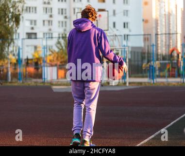 Adolescente carino in felpa con cappuccio viola che gioca a basket. Ragazzo giovane con palla imparare dribble e sparare sul campo della città. Hobby per bambini, Foto Stock