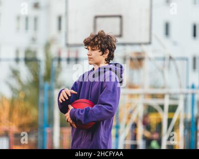 Adolescente carino in felpa con cappuccio viola che gioca a basket. Ragazzo giovane con palla imparare dribble e sparare sul campo della città. Hobby per bambini, Foto Stock