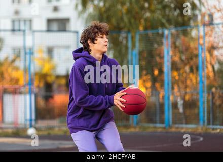 Adolescente carino in felpa con cappuccio viola che gioca a basket. Ragazzo giovane con palla imparare dribble e sparare sul campo della città. Hobby per bambini, Foto Stock