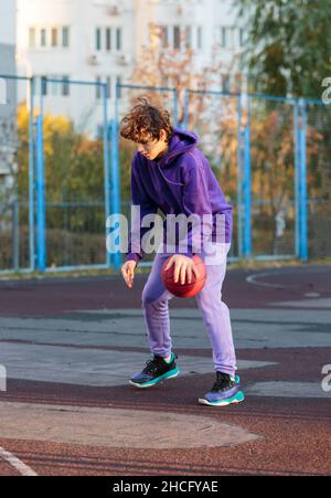 Adolescente carino in felpa con cappuccio viola che gioca a basket. Ragazzo giovane con palla imparare dribble e sparare sul campo della città. Hobby per bambini, Foto Stock