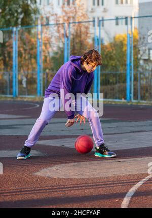 Adolescente carino in felpa con cappuccio viola che gioca a basket. Ragazzo giovane con palla imparare dribble e sparare sul campo della città. Hobby per bambini, Foto Stock