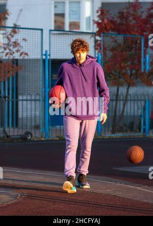 Adolescente carino in felpa con cappuccio viola che gioca a basket. Ragazzo giovane con palla imparare dribble e sparare sul campo della città. Hobby per bambini, Foto Stock