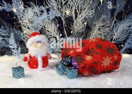 Un giocattolo a maglia Babbo Natale si erge sulla neve artificiale e una borsa di lino rosso con regali si trova accanto ad essa sullo sfondo di blu e bianco artificiale t Foto Stock