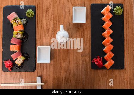 vista dall'alto di piatti neri con deliziosi sushi rotolini vicino a bacchette, pentola di salsa di soia e ciotola su tavola di legno Foto Stock