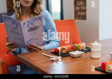 vista ritagliata del menu di lettura della donna sfocata vicino a gustosi sushi rotolini sul tavolo Foto Stock