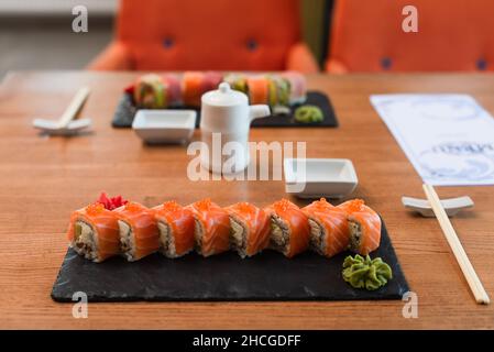 piatti neri con gustosi sushi rotolini vicino a bacchette, pentola di salsa di soia e menu sfocato su tavola di legno Foto Stock
