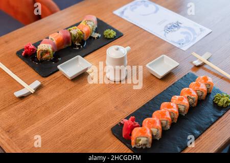 piatti neri con deliziosi panini di sushi con wasabi e zenzero vicino al piatto di salsa di soia e opuscolo del menu su tavola di legno Foto Stock