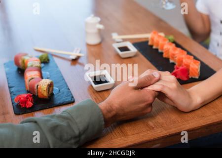 vista croppata della coppia che tiene le mani vicino ai rotoli di sushi e alle ciotole con salsa di soia sul tavolo Foto Stock