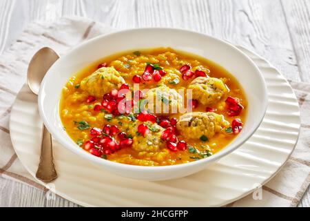 Primo piano di Anar di Ash-e, zuppa invernale di melagrana giallo persiano con polpette, zuppa spessa, in una ciotola bianca su un tavolo di legno bianco testurizzato Foto Stock