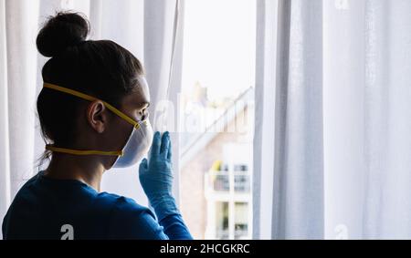 Annoiato donna in quarantena corona con maschera di protezione FFP2 guardando fuori dalla finestra per la strada Foto Stock